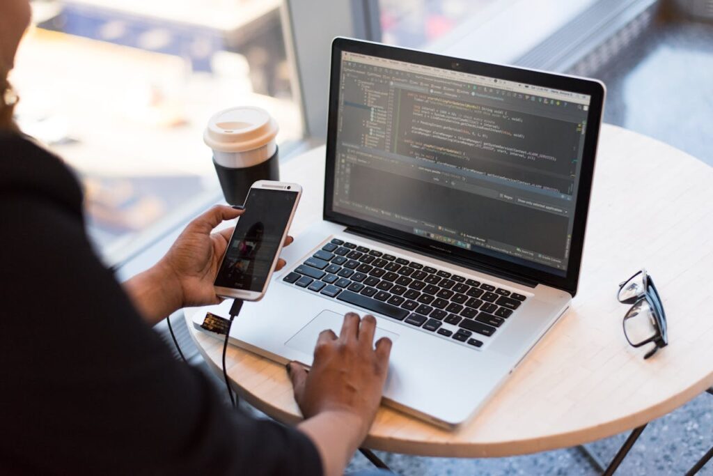 pexels-photo-1181244 Developer working remotely, coding on a laptop with phone in hand, showcasing modern work culture.